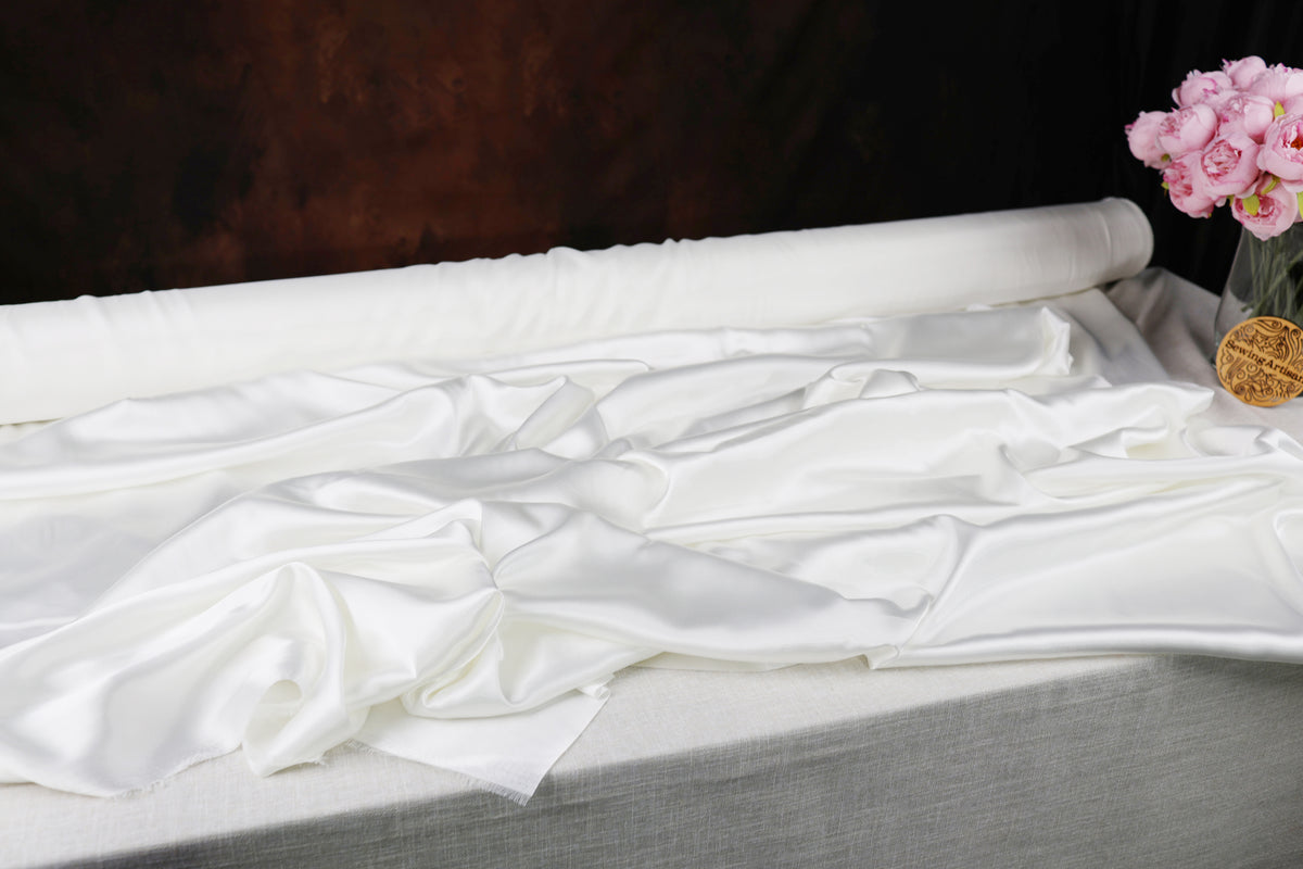 a white fabric, possibly silk or satin, lying on a surface, with a vase of pink flowers placed next to it.