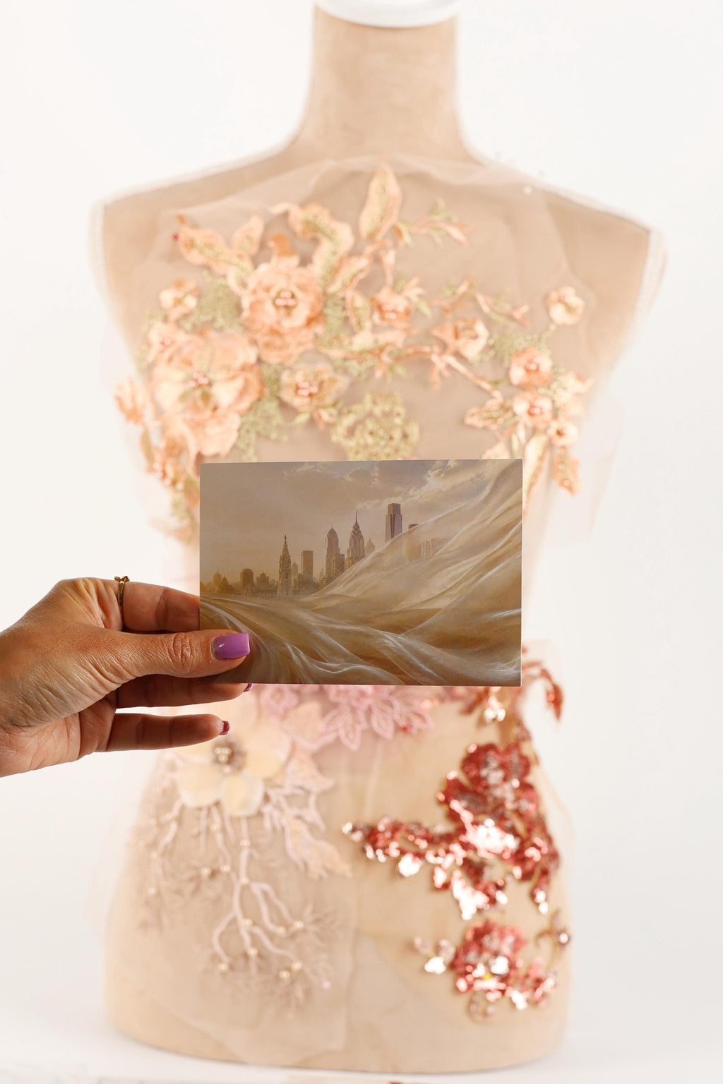 A hand holding a photograph of a cityscape, with a dress featuring a floral pattern and sequins on the bust.