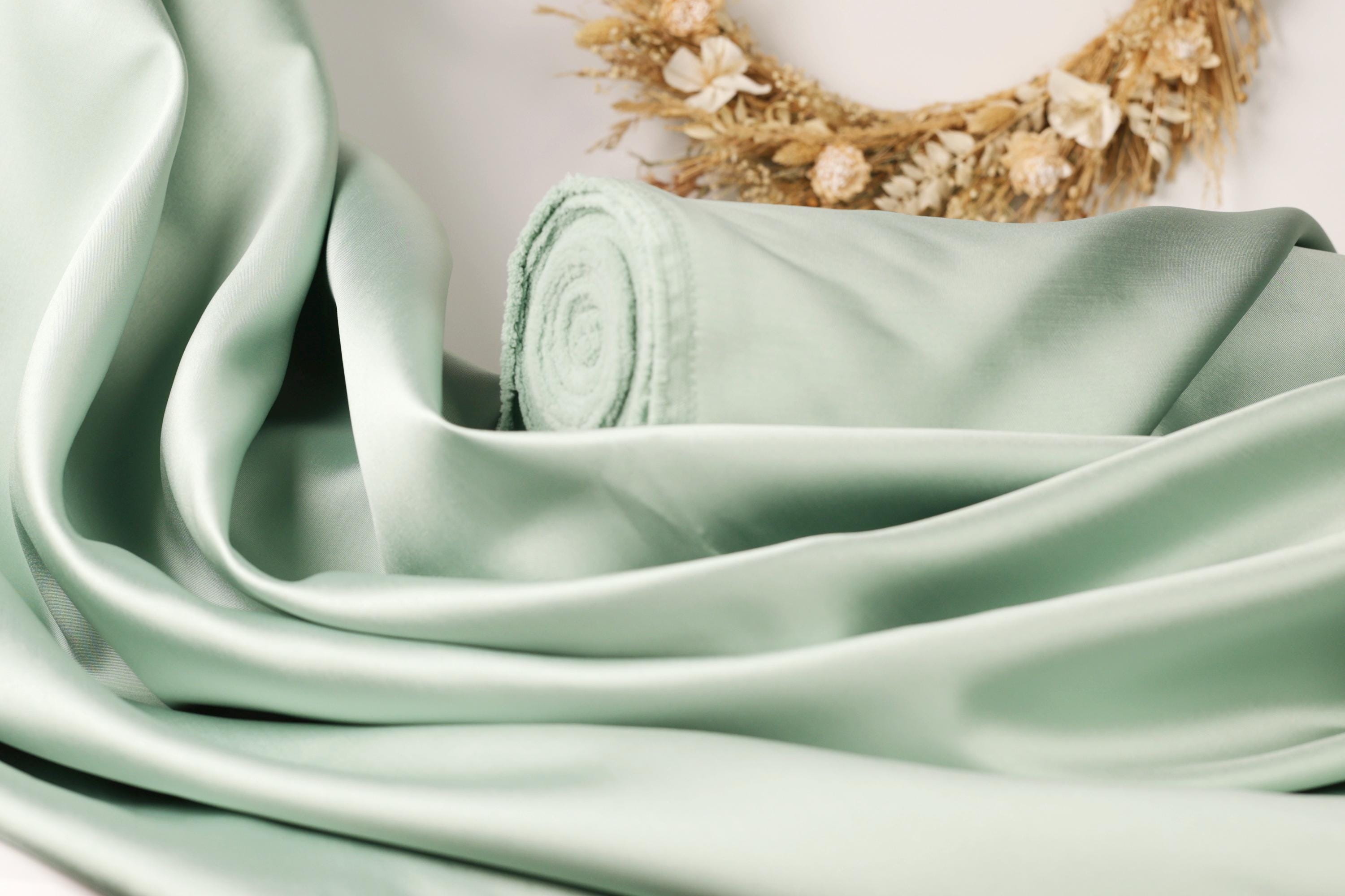 A close-up view of a light green satin fabric, possibly silk, with a rolled-up end visible. In the background, there is a wreath made of dried flowers.