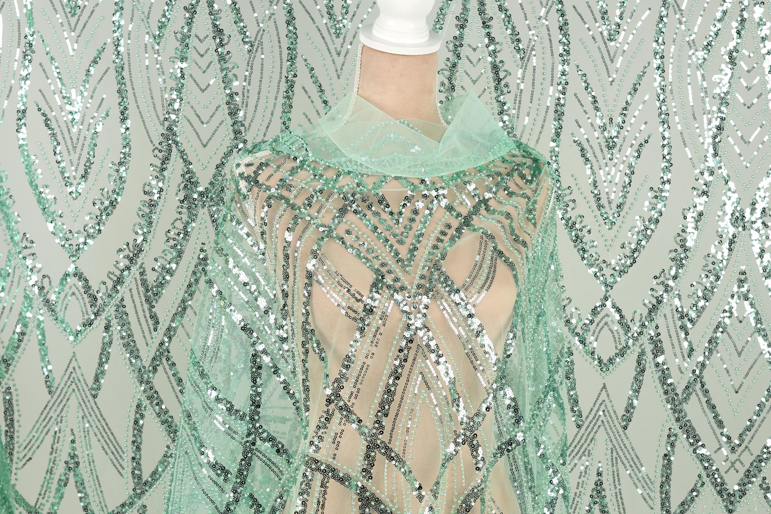 a mannequin wearing a green sequin dress with a sheer overlay. The dress has a pattern of green and silver sequins, creating a visually striking and intricate design.