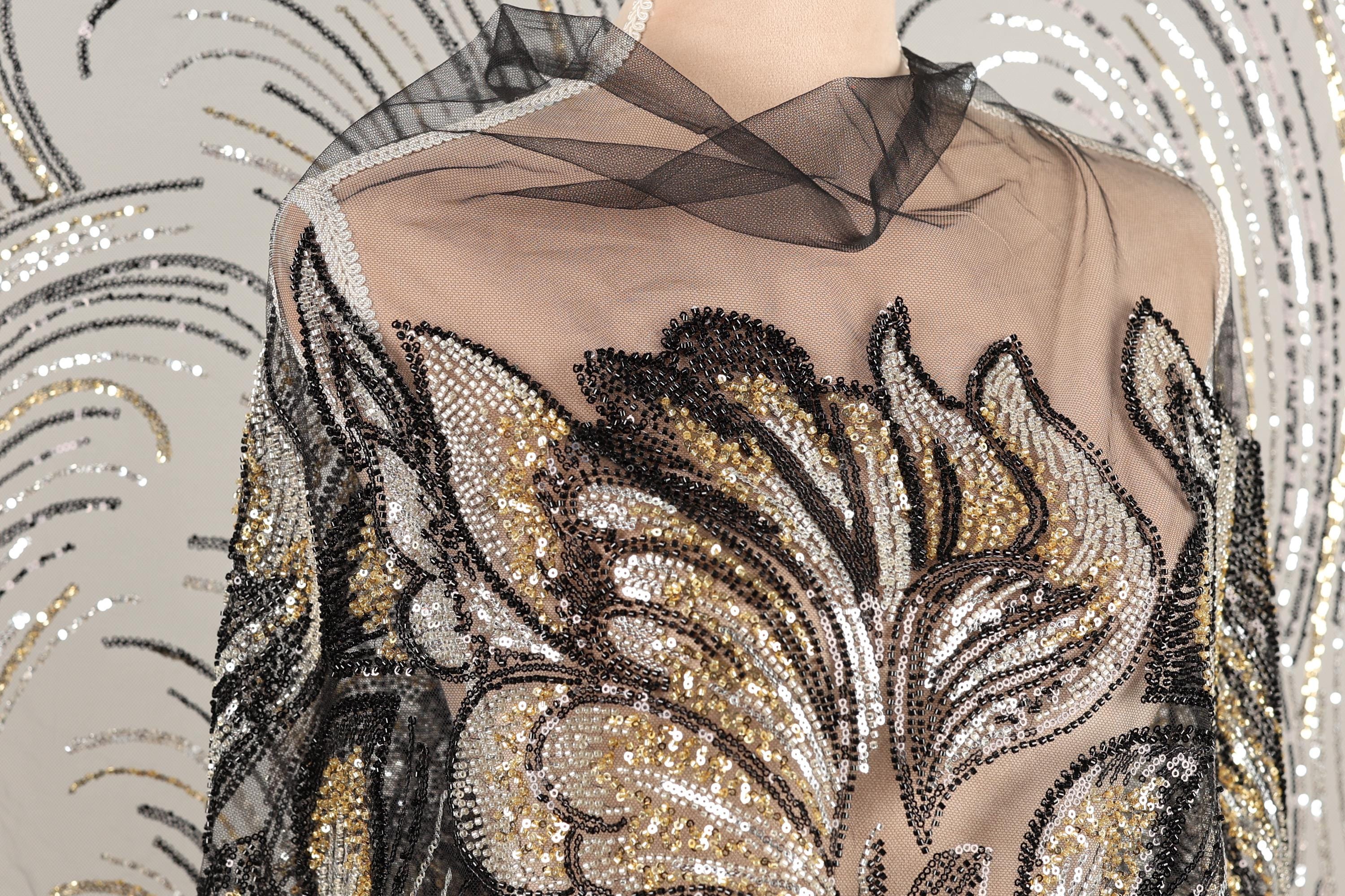 a mannequin wearing a dress with a sheer, sheer-like fabric and a floral pattern. The dress is adorned with sequins and beads, giving it a glamorous and elegant appearance.