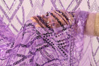 A hand is holding a piece of sequin fabric with a purple and black pattern, against a background of a white wall with a geometric pattern.