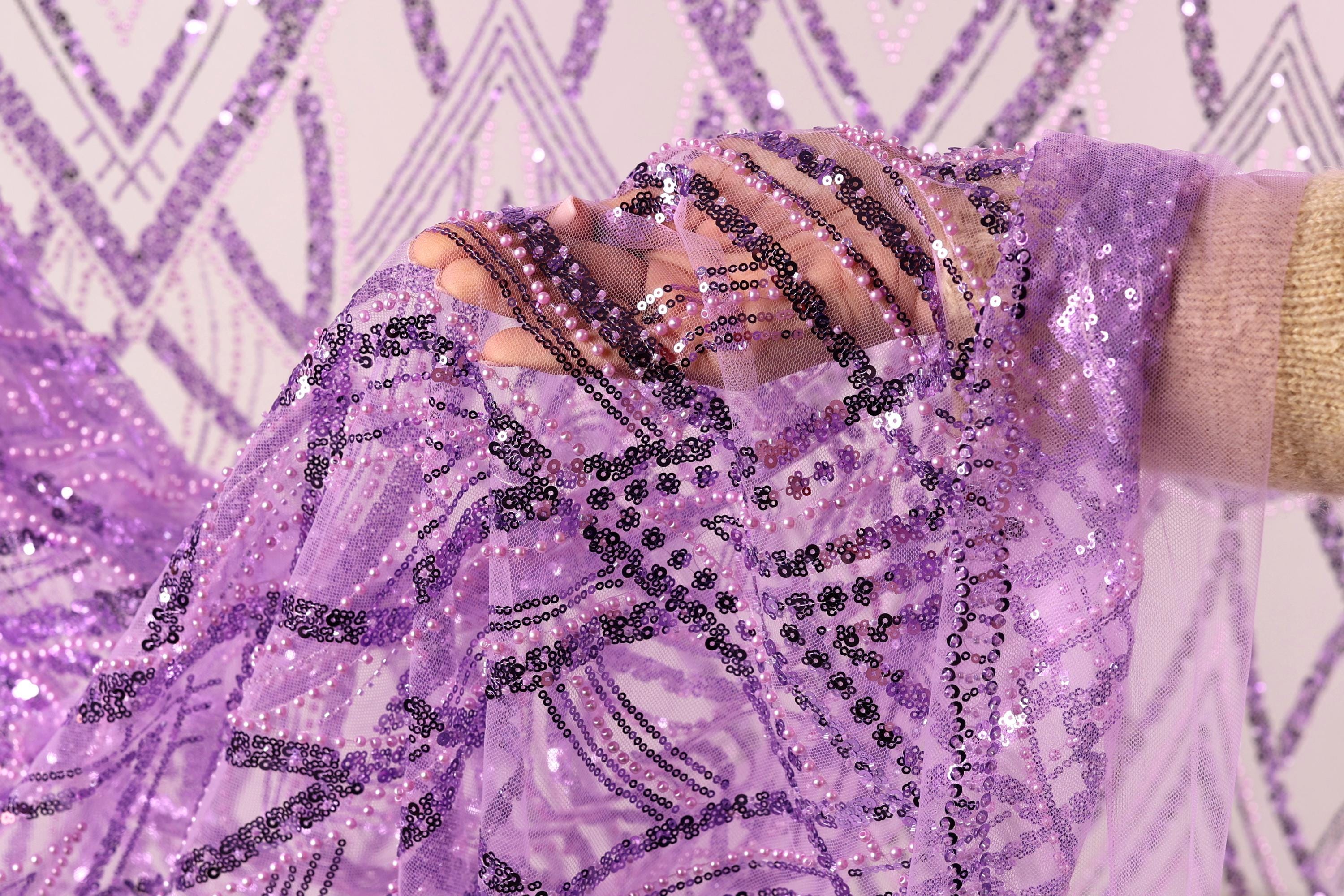 A hand is holding a piece of sequin fabric with a purple and black pattern, against a background of a white wall with a geometric pattern.