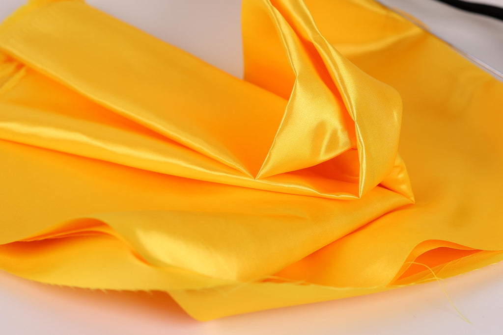 a close-up view of a yellow fabric, possibly silk or satin, with a soft, shiny texture. The fabric appears to be folded or crumpled, creating a sense of movement and texture.