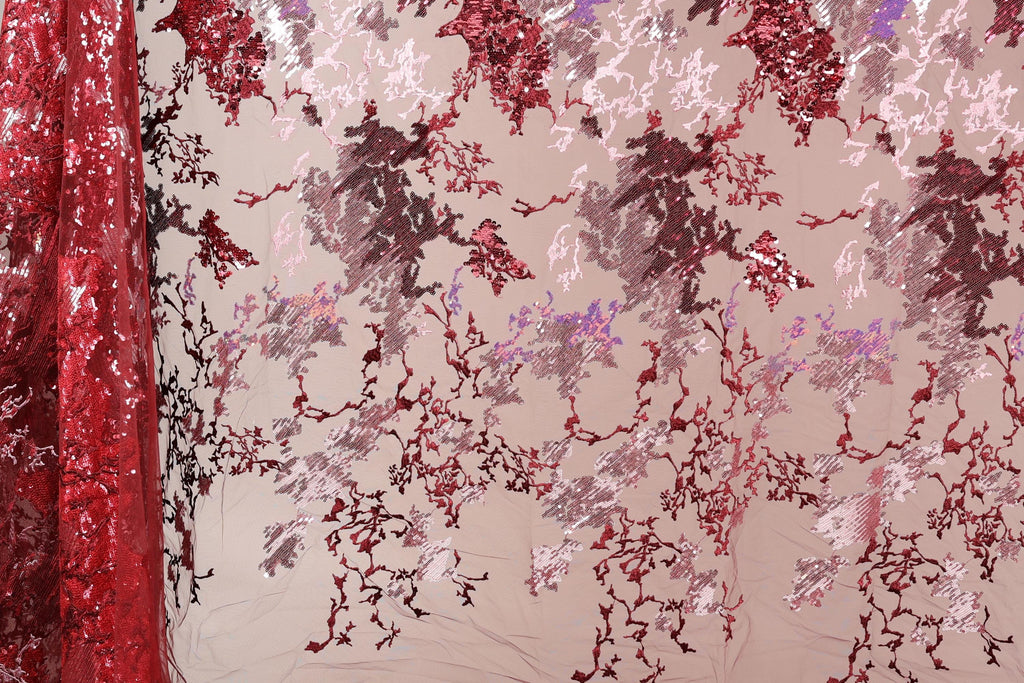 a close-up view of a fabric with a floral pattern in shades of red and pink. The fabric appears to be made of sequin material, creating a visually striking and intricate design.