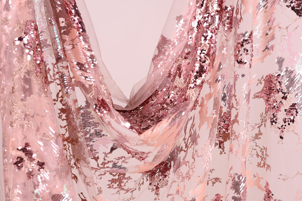 a close-up view of a fabric with a pink and white sequined pattern. The fabric appears to be a sheer material, with a delicate and intricate design. The pattern consists of small sequins and beads that create a visually striking and eye-catching appearance. The fabric is draped in a way that it cascades down, creating a sense of movement and elegance.