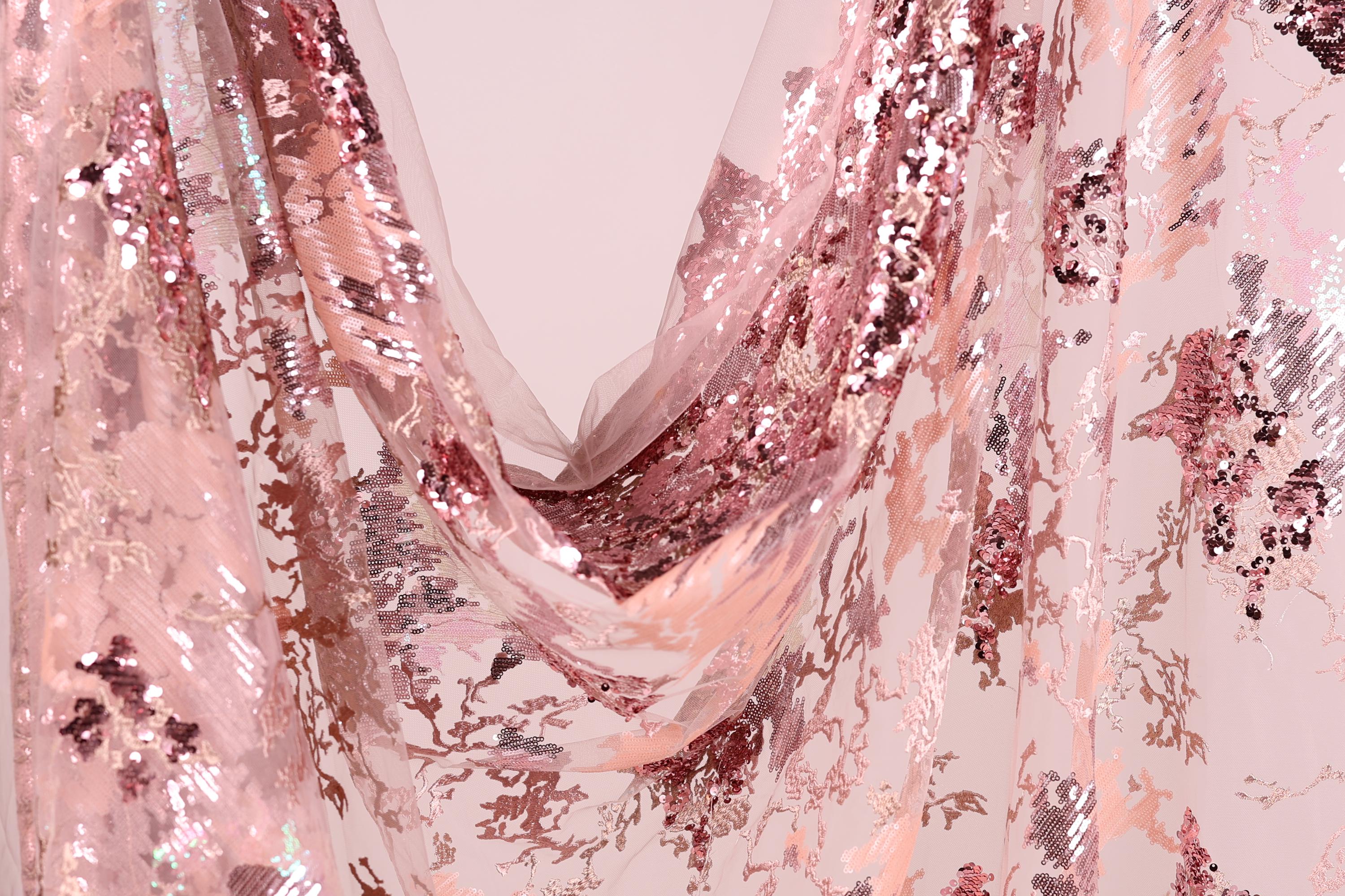 a close-up view of a fabric with a pink and white sequined pattern. The fabric appears to be a sheer material, with a delicate and intricate design. The pattern consists of small sequins and beads that create a visually striking and eye-catching appearance. The fabric is draped in a way that it cascades down, creating a sense of movement and elegance.