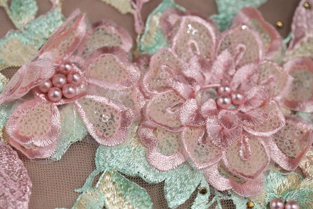 The image is a close-up of a delicate, intricately embroidered fabric with pink and green floral elements. The fabric features a variety of pink flowers, including a large, fully bloomed flower in the center, surrounded by smaller flowers and leaves. The fabric appears to be made of a soft, delicate material, such as lace or chiffon, and is adorned with small beads or sequins that add texture and detail to the design.