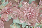 The image is a close-up of a delicate, intricately embroidered fabric with pink and green floral elements. The fabric features a variety of pink flowers, including a large, fully bloomed flower in the center, surrounded by smaller flowers and leaves. The fabric appears to be made of a soft, delicate material, such as lace or chiffon, and is adorned with small beads or sequins that add texture and detail to the design.