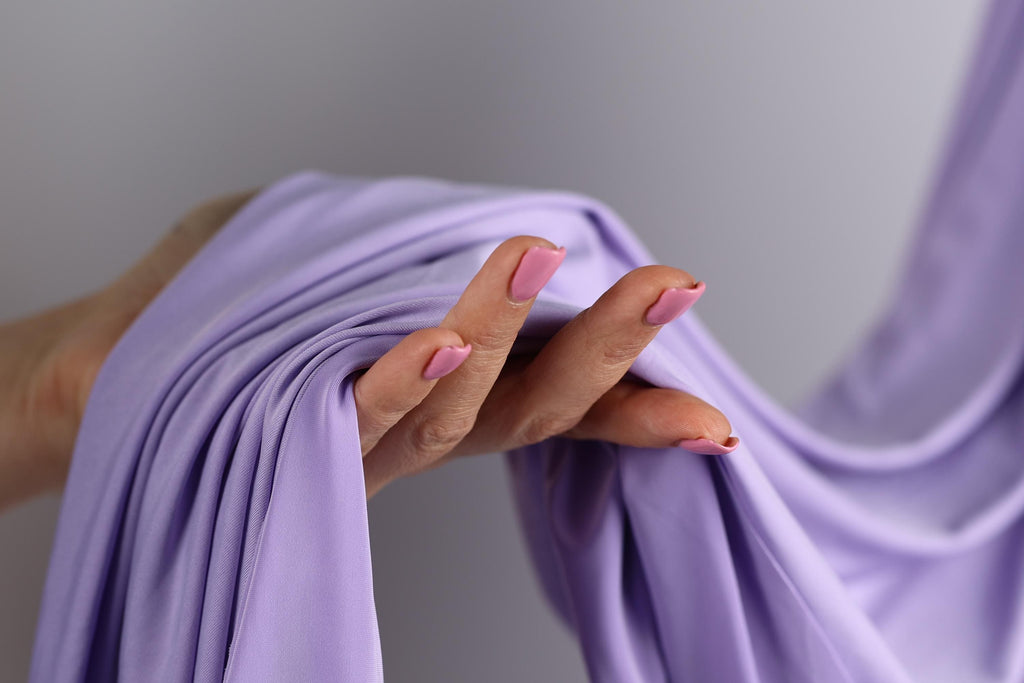 A hand with pink nail polish is holding a purple fabric, with the fabric partially rolled up.