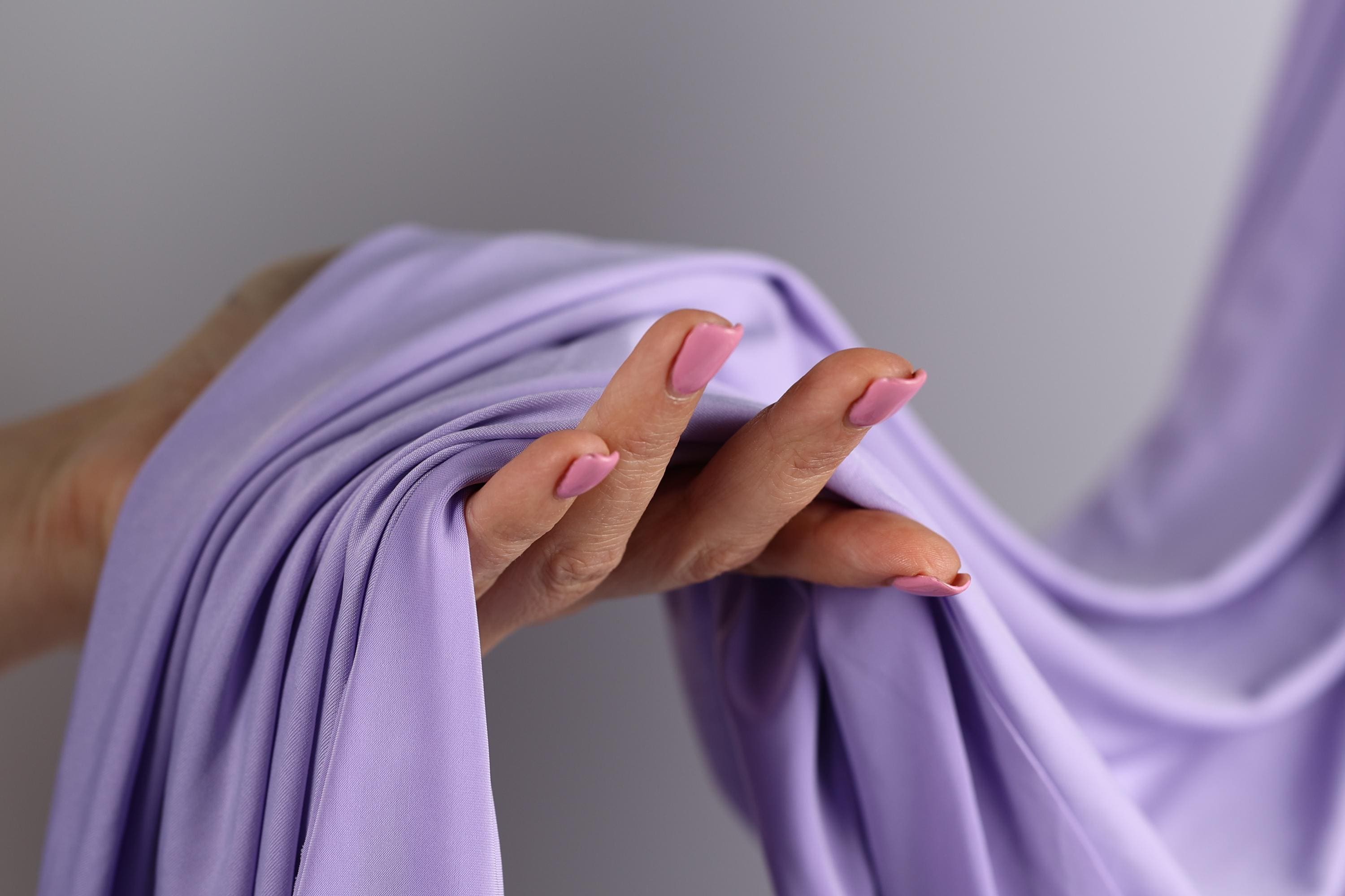 A hand with pink nail polish is holding a purple fabric, with the fabric partially rolled up.