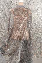 a mannequin wearing a shimmering, sequined dress with a floral pattern. The dress has a sheer, flowing design that creates a sense of movement and elegance. The background is a light gray color, which contrasts with the vibrant colors of the sequinwork and the dress.
