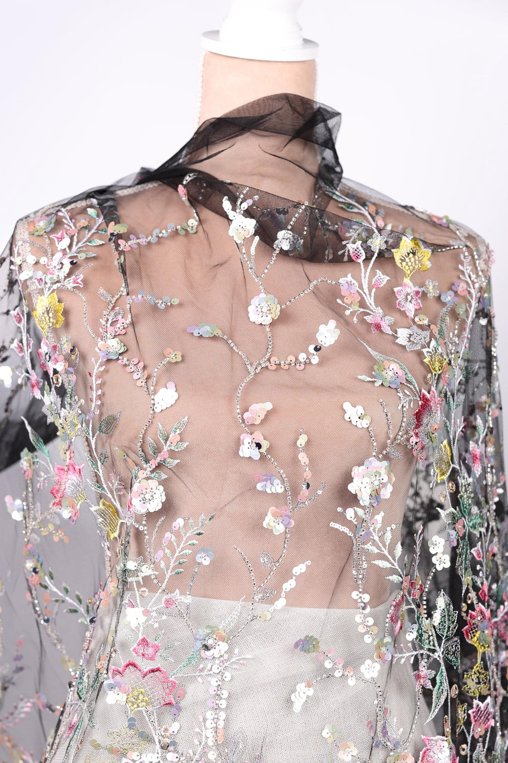 a mannequin wearing a sheer, floral-patterned fabric with a black collar. The fabric is adorned with a variety of colorful flowers and leaves, creating a visually striking and intricate design.
