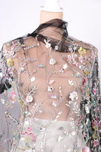 a mannequin wearing a sheer, floral-patterned fabric with a black collar. The fabric is adorned with a variety of colorful flowers and leaves, creating a visually striking and intricate design.