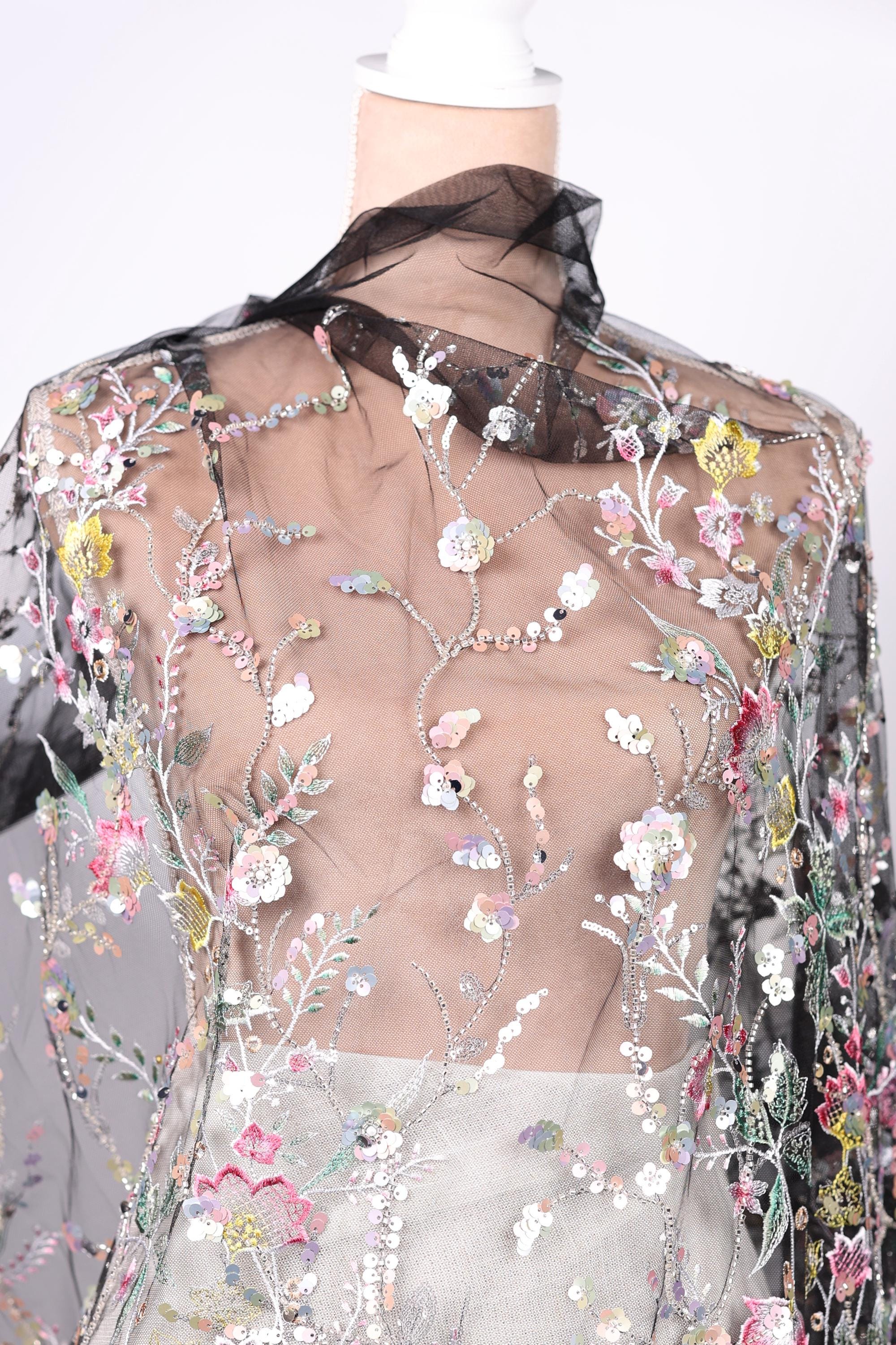 a mannequin wearing a sheer, floral-patterned fabric with a black collar. The fabric is adorned with a variety of colorful flowers and leaves, creating a visually striking and intricate design.