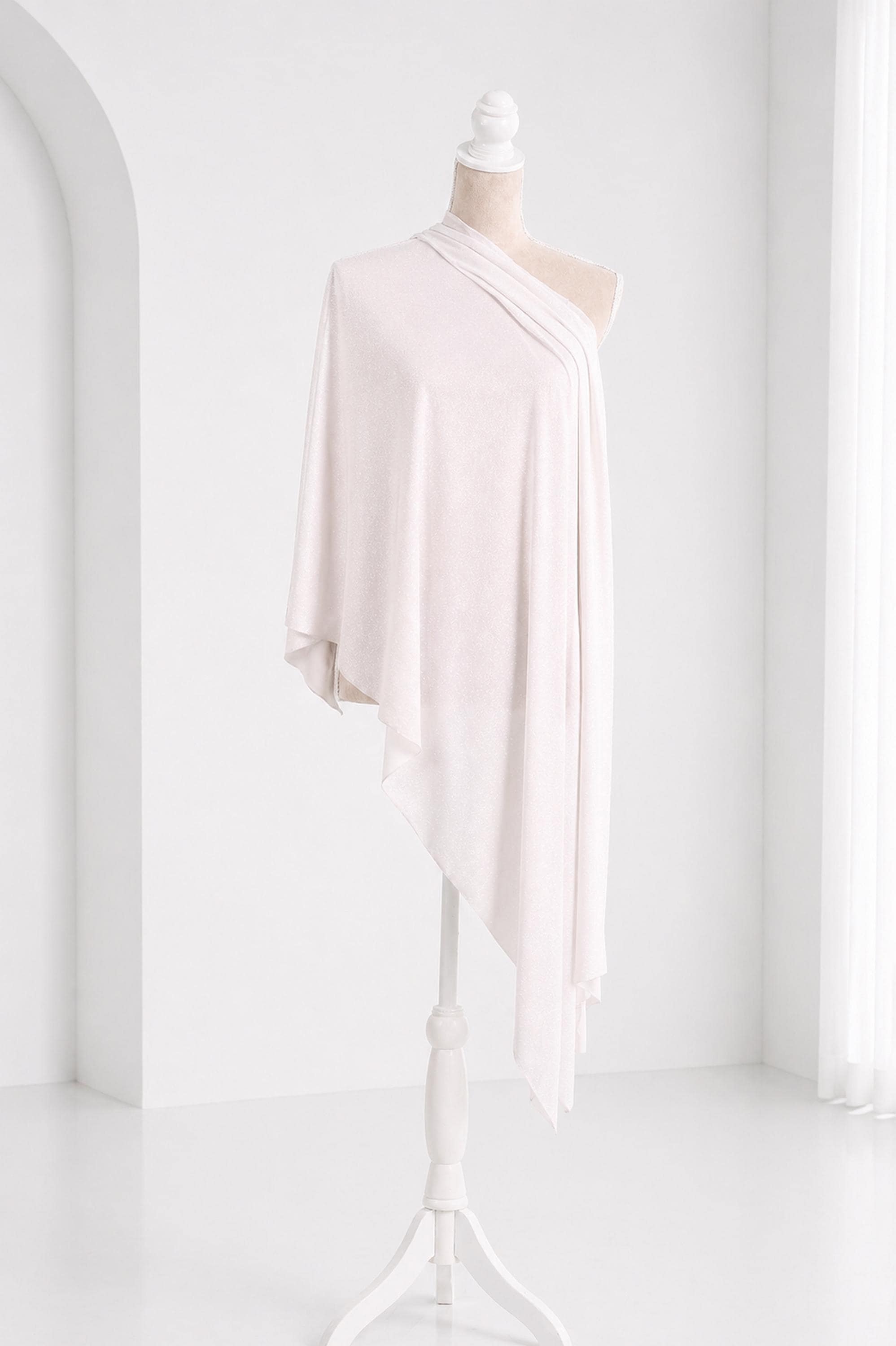 A white mannequin wearing a draped, flowing white garment stands on a white stand against a white background.