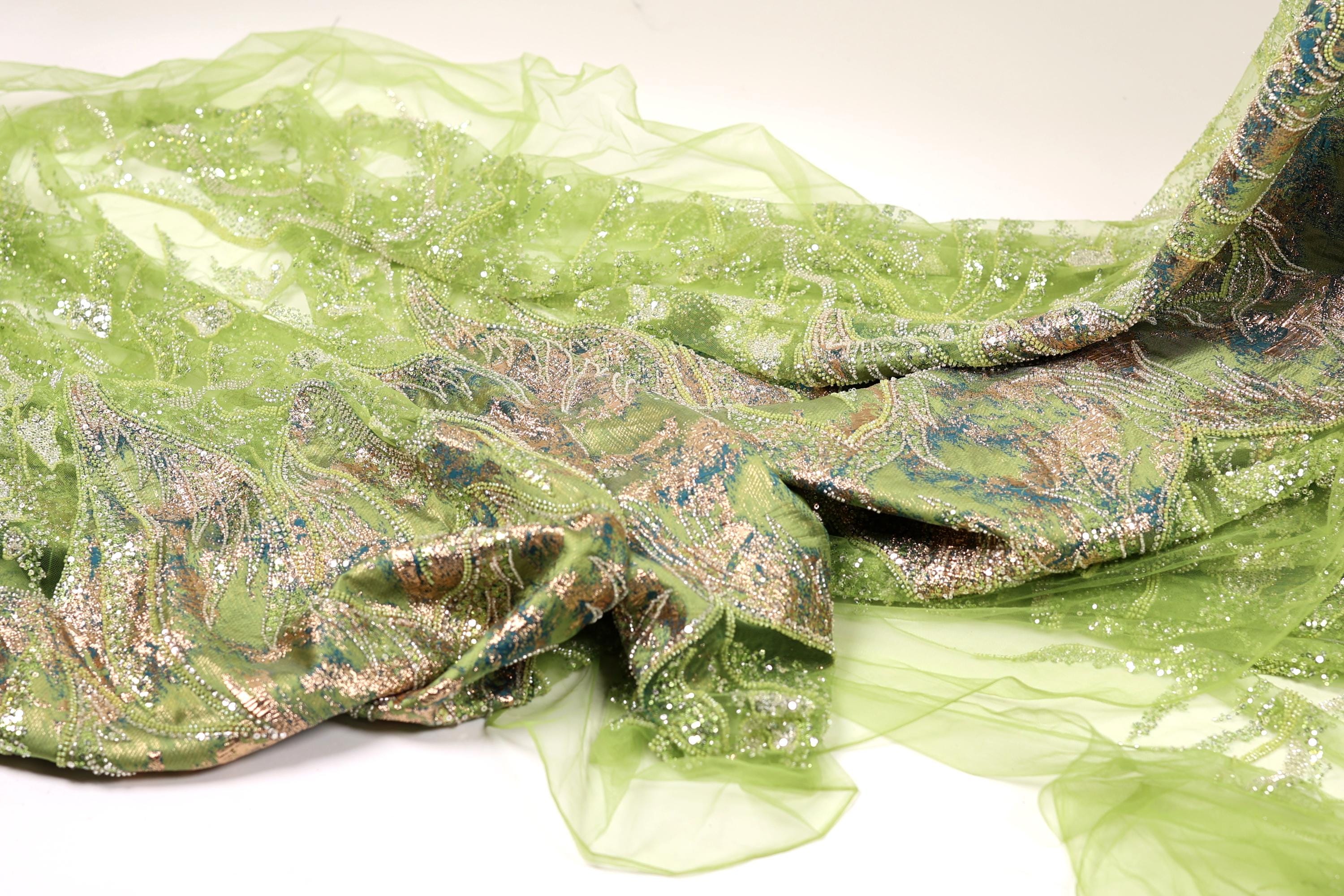 a close-up view of a green, shimmering fabric with a floral pattern. The fabric appears to be made of a sheer material, giving it a delicate and airy appearance. The intricate design and vibrant colors of the fabric create a visually striking contrast against the plain white background.