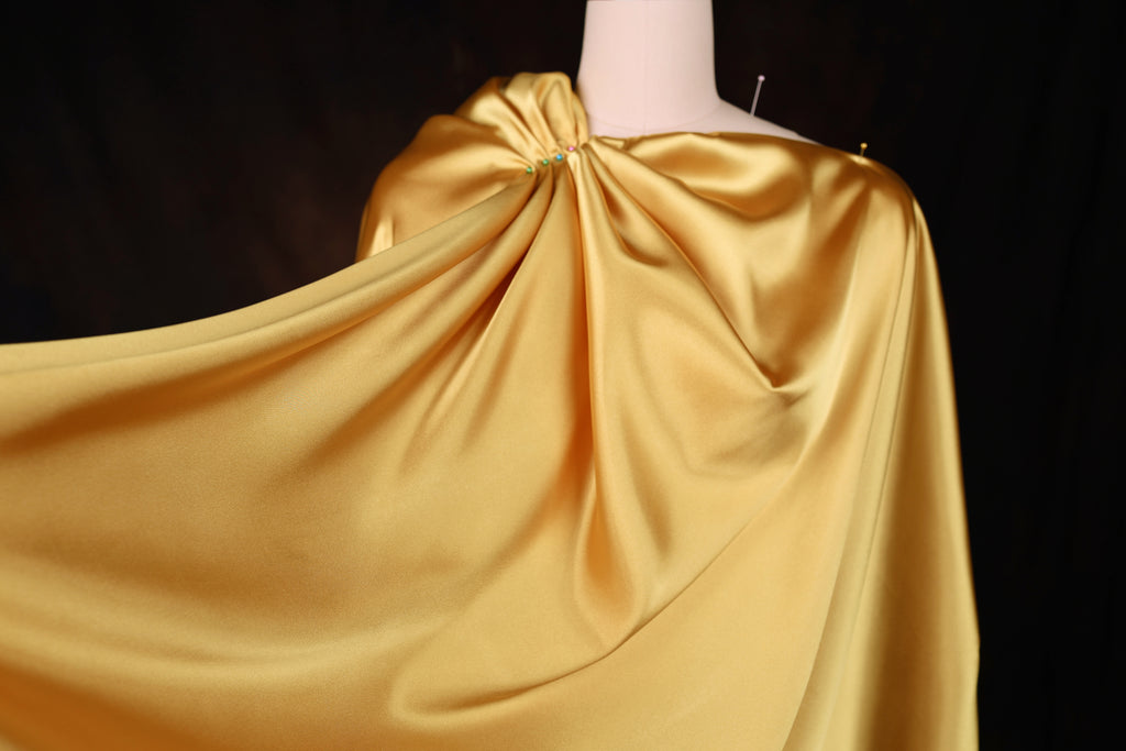 A close-up view of a gold satin cape draped over a mannequin, with the fabric flowing and creating a draped effect.
