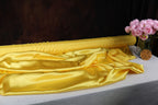a roll of shiny yellow satin fabric and a vase of pink flowers on a table.