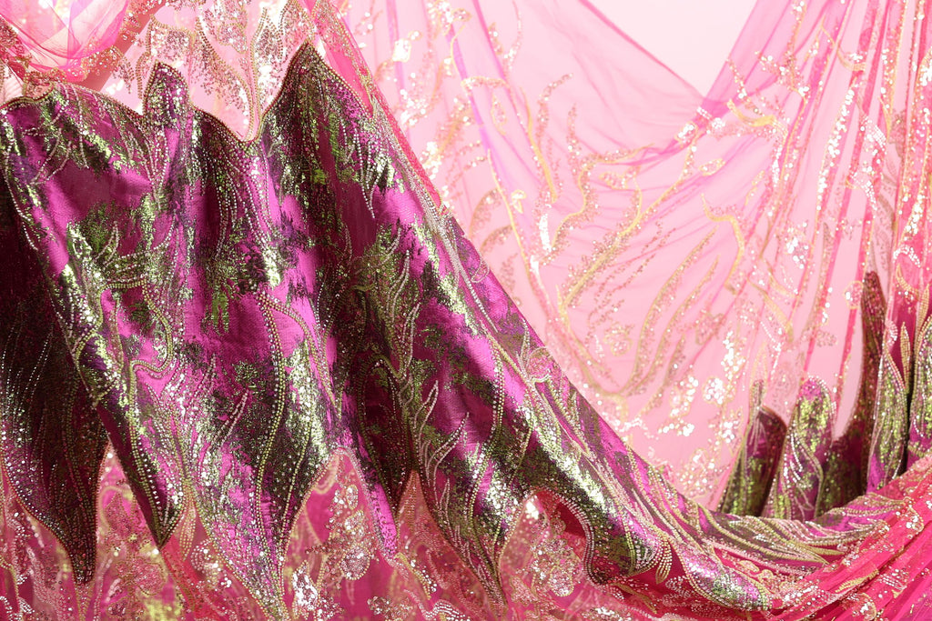 a close-up view of a fabric with a pink and green floral pattern. The fabric appears to be made of a shiny, sequined material, giving it a luxurious and eye-catching appearance.