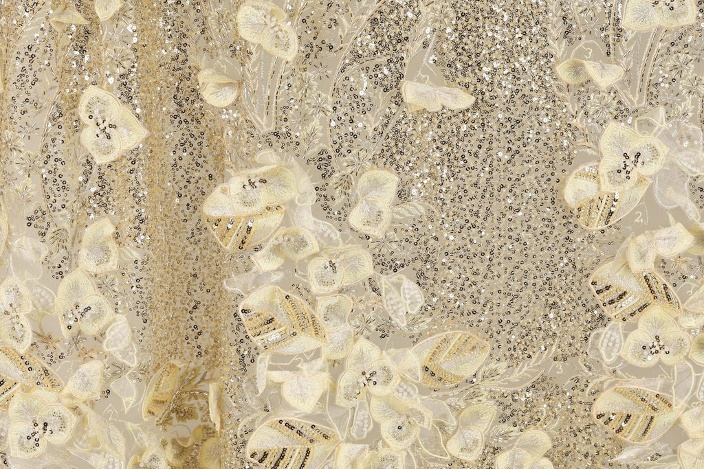 The image is a close-up of a fabric with a gold and white floral pattern, featuring small sequins and beads.