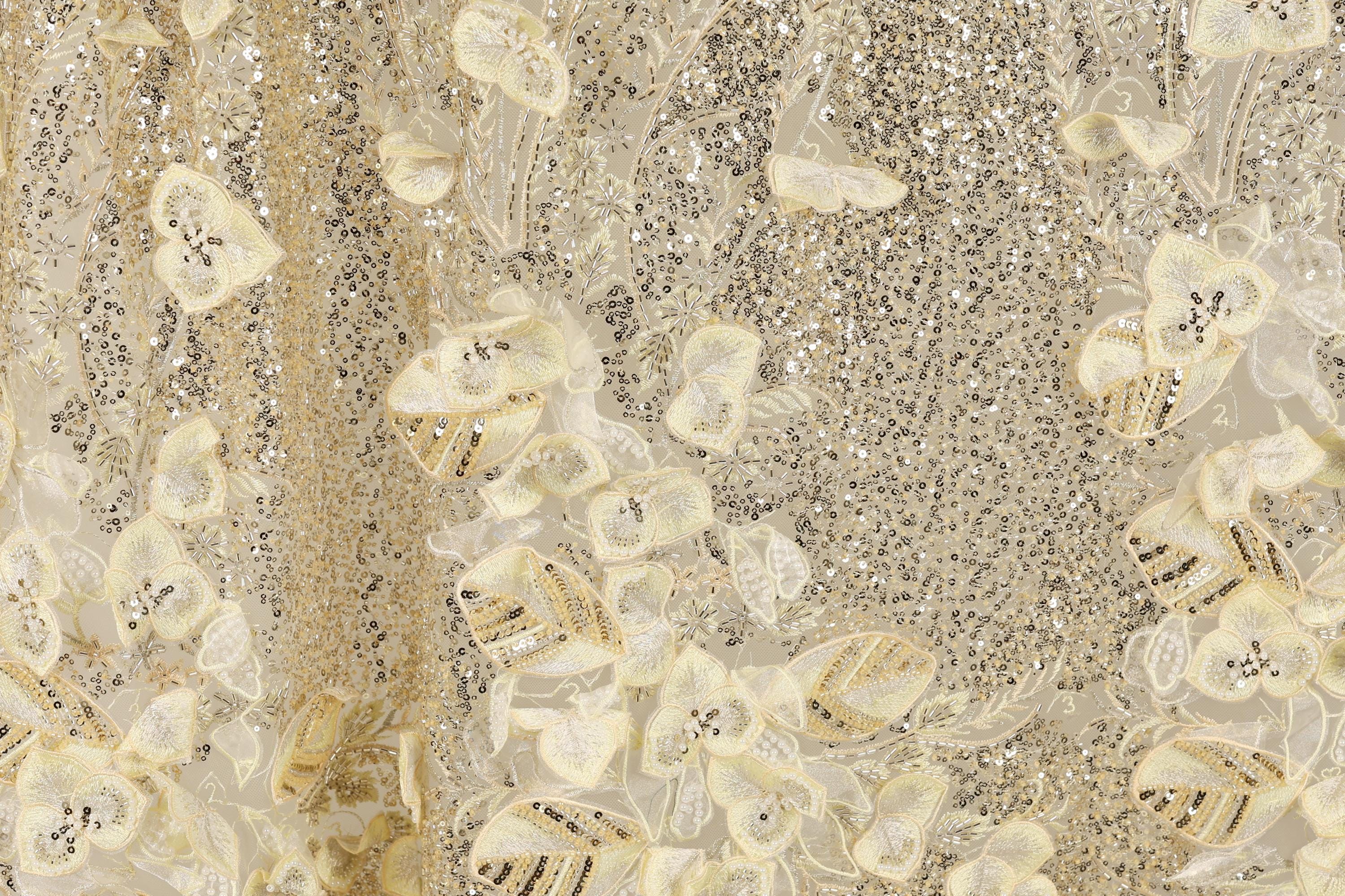 The image is a close-up of a fabric with a gold and white floral pattern, featuring small sequins and beads.