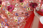 A hand is holding a red fabric with a floral pattern of various colorful flowers, including red, pink, yellow, and purple flowers, against a pink background.