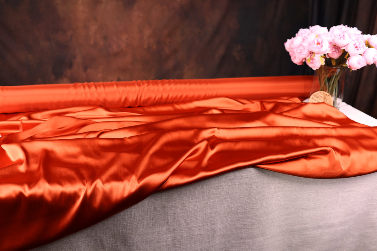 a large roll of orange satin fabric, a vase of pink flowers, and a gray tablecloth.