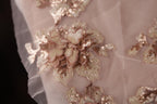a close-up view of a delicate, pink-colored fabric with intricate gold and white sequin details. The fabric appears to be a lace or sheer material, with the sequins adding a touch of sparkle and elegance to the overall design.