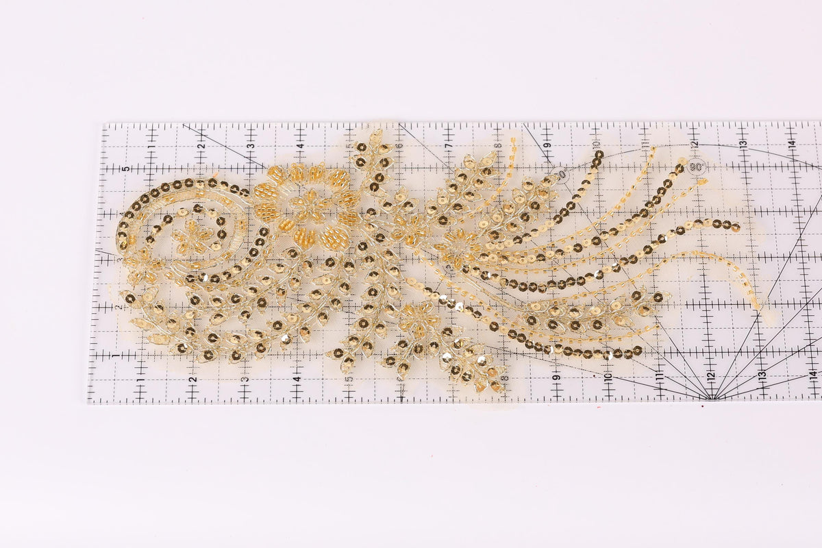 a decorative piece with a gold and black color scheme, including a floral pattern and a swirling design. The piece appears to be a handmade embroidery or beading work, with a ruler visible in the background.