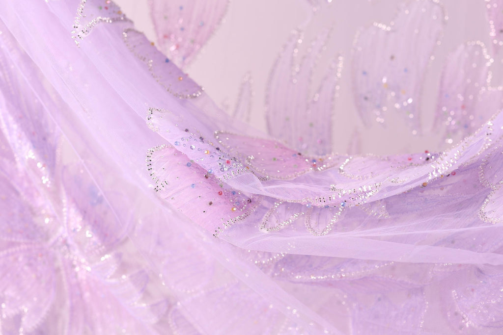 A close-up view of a delicate, flowing fabric with a soft, pastel pink hue. The fabric appears to be made of sheer material, with small, glittery embellishments scattered across its surface, adding a touch of sparkle and elegance to the overall design.