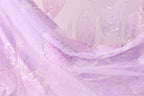 A close-up view of a delicate, flowing fabric with a soft, pastel pink hue. The fabric appears to be made of sheer material, with small, glittery embellishments scattered across its surface, adding a touch of sparkle and elegance to the overall design.