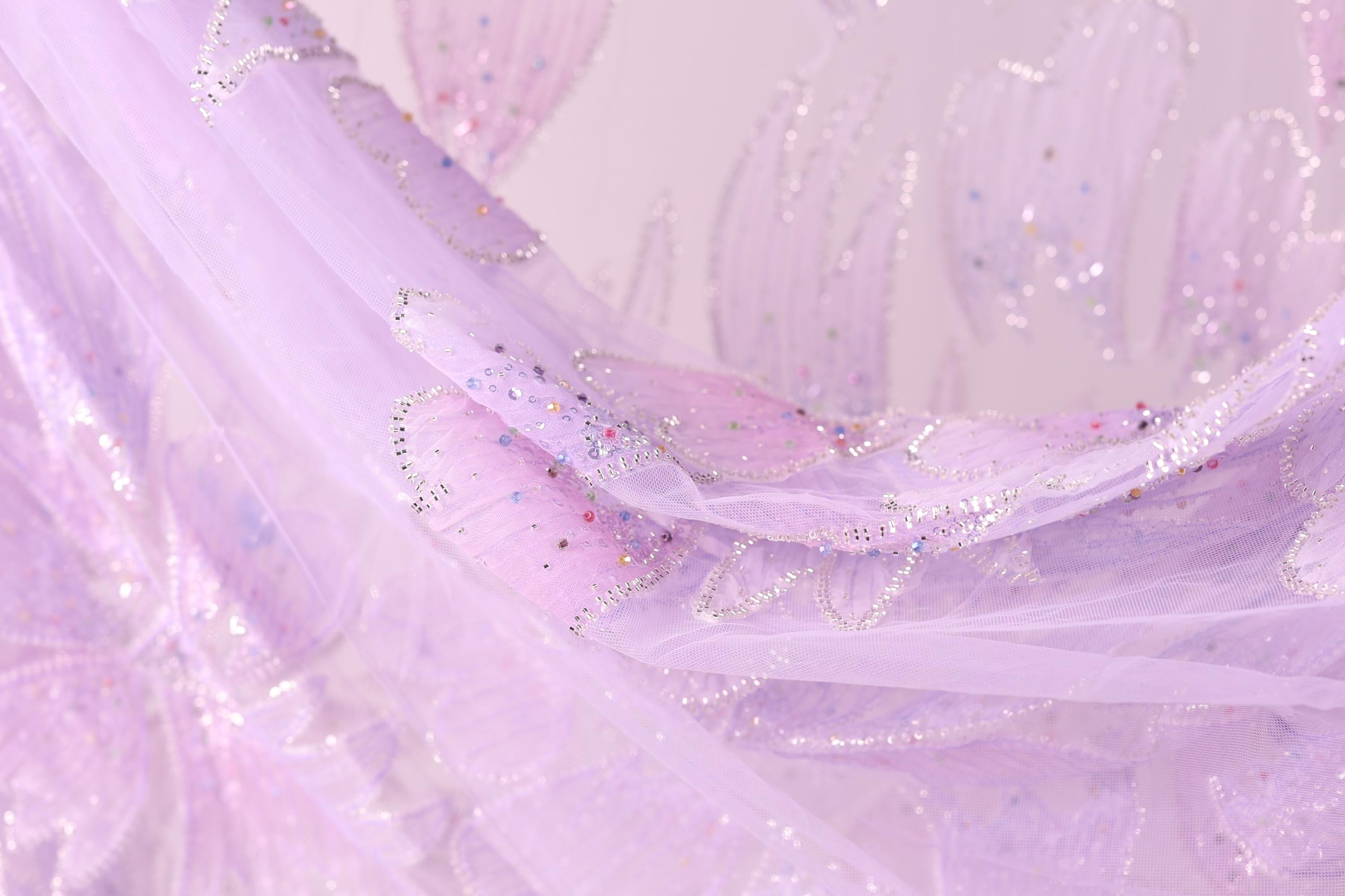A close-up view of a delicate, flowing fabric with a soft, pastel pink hue. The fabric appears to be made of sheer material, with small, glittery embellishments scattered across its surface, adding a touch of sparkle and elegance to the overall design.