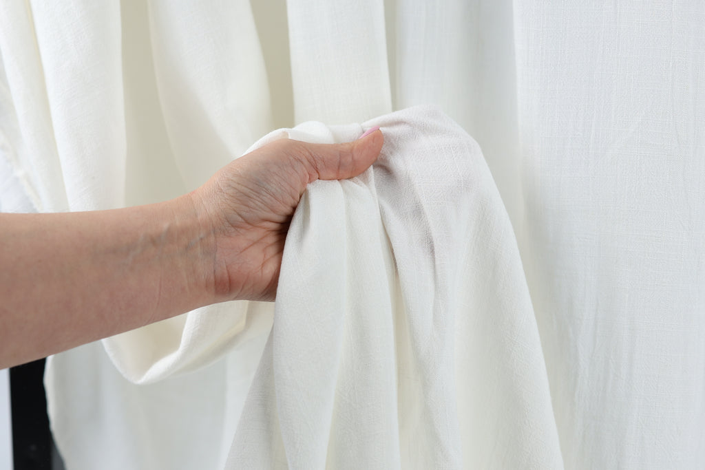 A hand is holding a white fabric, possibly a curtain or sheet, in front of a white background.