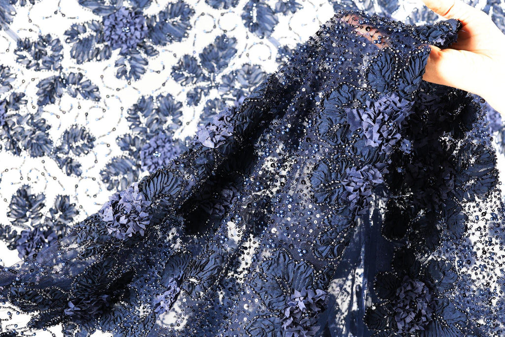 A close-up view of a fabric with a floral pattern, featuring a mix of blue and white colors. The fabric appears to be made of lace or a similar type of material, with intricate details and a textured appearance.