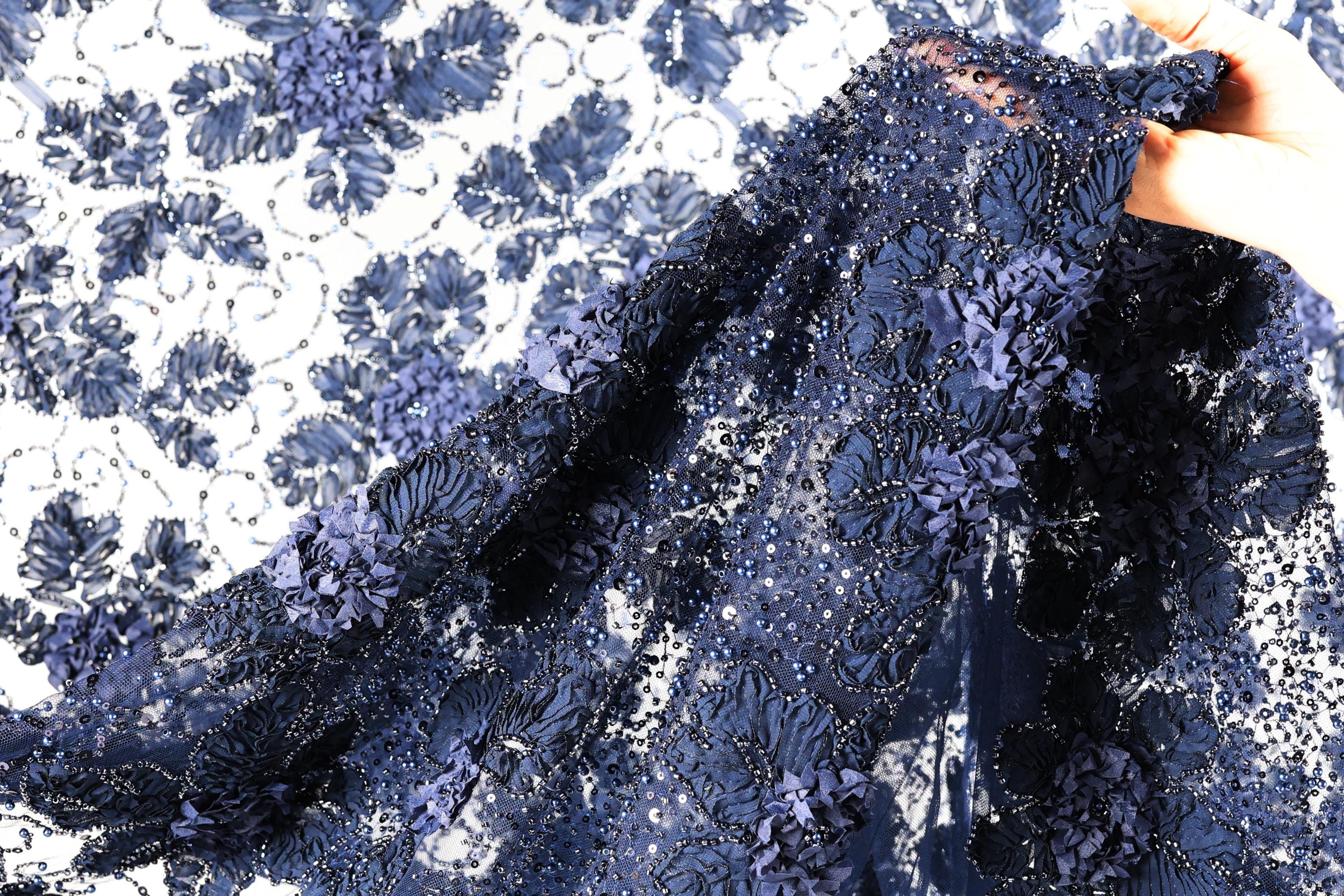 A close-up view of a fabric with a floral pattern, featuring a mix of blue and white colors. The fabric appears to be made of lace or a similar type of material, with intricate details and a textured appearance.