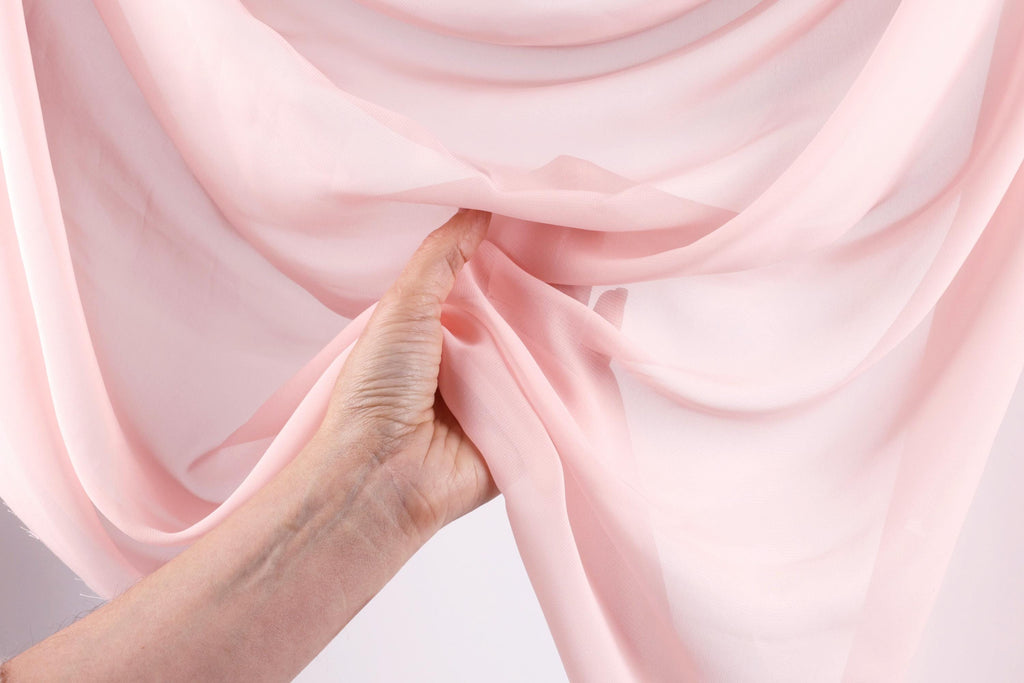 A hand is holding a pink fabric, possibly silk or chiffon, against a white background.