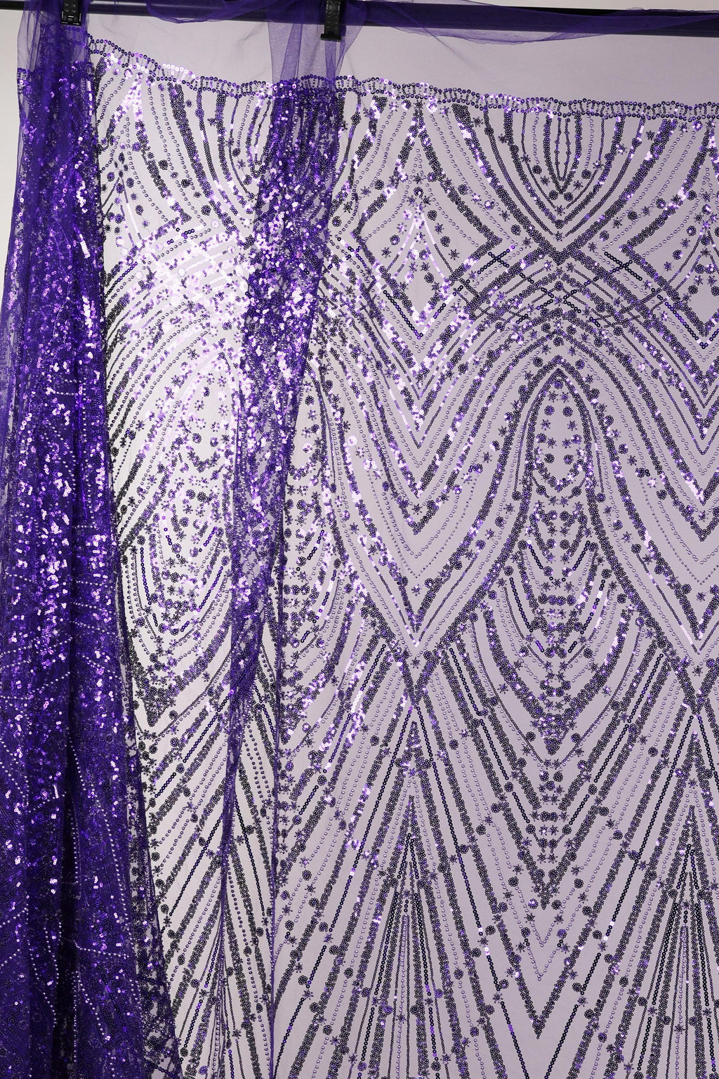 a close-up view of a sequin fabric with a purple and white pattern. The fabric appears to be made of a shiny, metallic material and has a repeating pattern of lines and dots in shades of purple and white. The fabric is hanging on a rod, and the background is a plain white wall.
