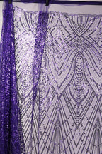 a close-up view of a sequin fabric with a purple and white pattern. The fabric appears to be made of a shiny, metallic material and has a repeating pattern of lines and dots in shades of purple and white. The fabric is hanging on a rod, and the background is a plain white wall.