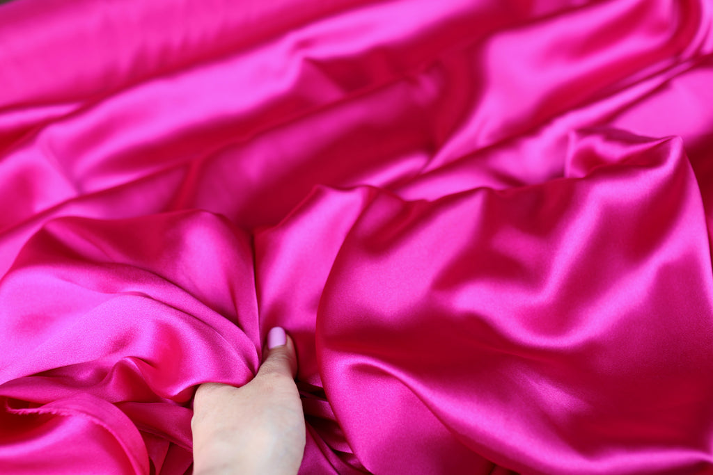A hand is holding a piece of bright pink satin fabric, which appears to be soft and smooth.