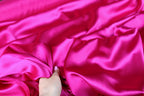 A hand is holding a piece of bright pink satin fabric, which appears to be soft and smooth.
