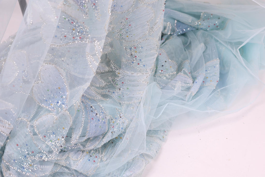 a close-up view of a light blue, sheer fabric with a floral pattern and small, multicolored sequins or beads scattered across it.