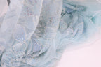 a close-up view of a light blue, sheer fabric with a floral pattern and small, multicolored sequins or beads scattered across it.