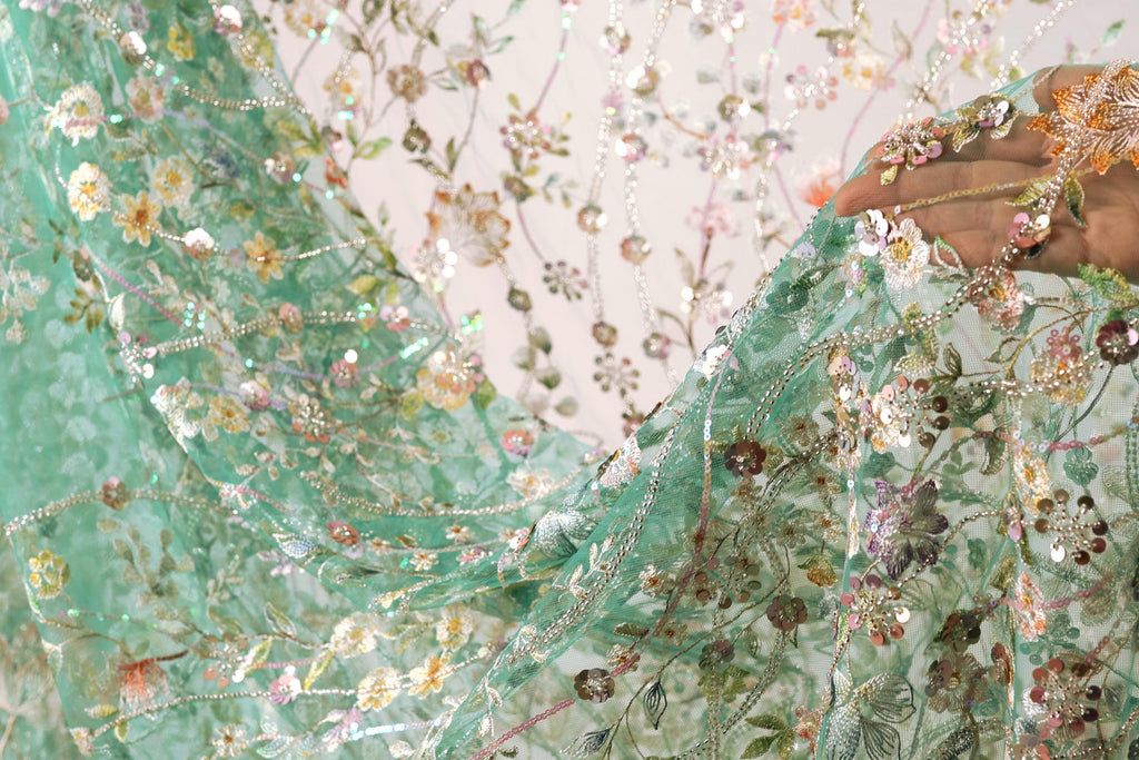 a close-up view of a hand holding a piece of fabric with a floral pattern. The fabric appears to be a light green material with a variety of colorful flowers and leaves, creating a visually appealing and intricate design.