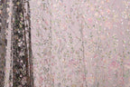 a close-up view of a fabric with a floral pattern, including pink and green flowers, as well as leaves and branches. The fabric appears to be made of a sheer material, giving it a delicate and airy appearance. The background of the image is a light pink color, which complements the floral pattern on the fabric.