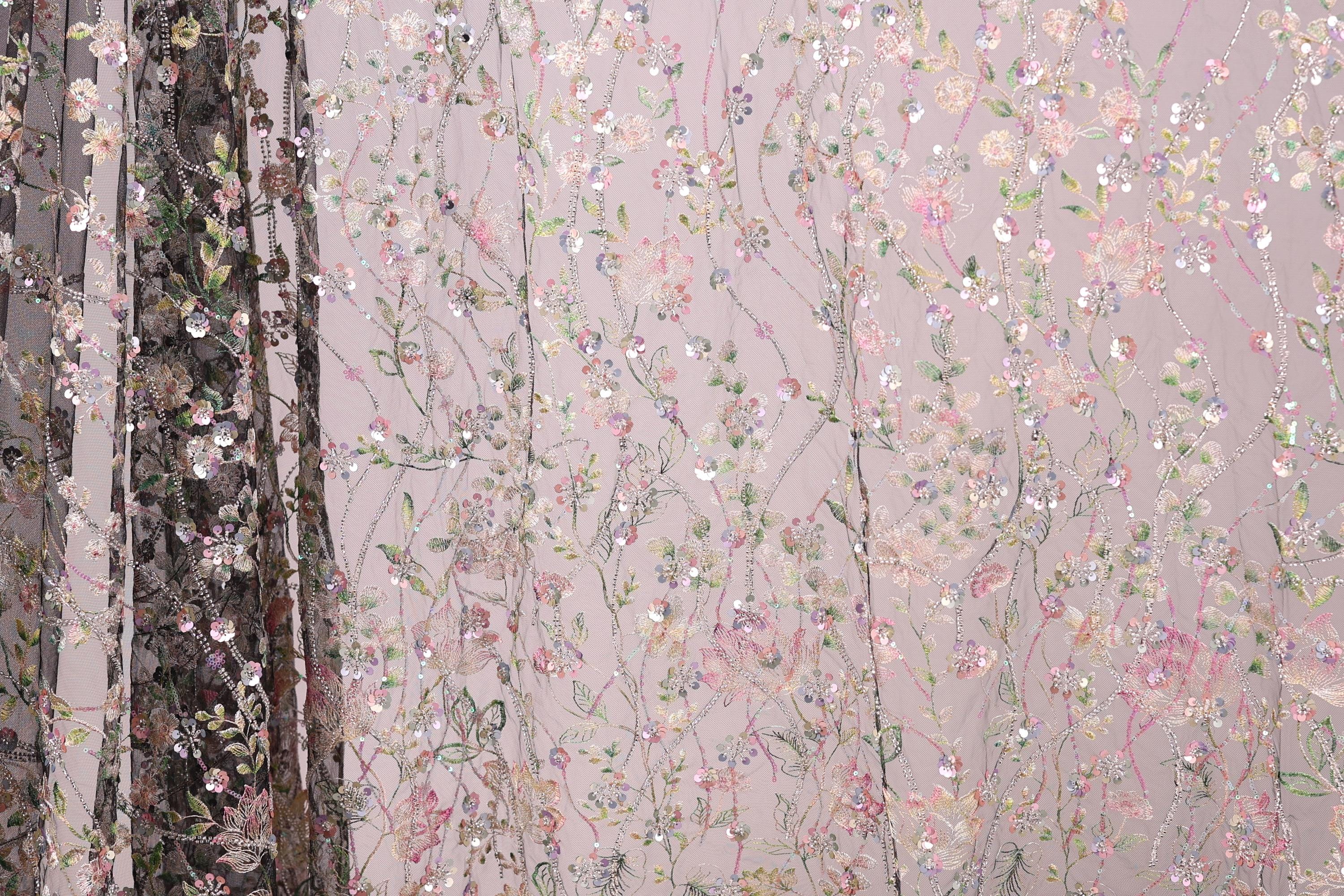 a close-up view of a fabric with a floral pattern, including pink and green flowers, as well as leaves and branches. The fabric appears to be made of a sheer material, giving it a delicate and airy appearance. The background of the image is a light pink color, which complements the floral pattern on the fabric.