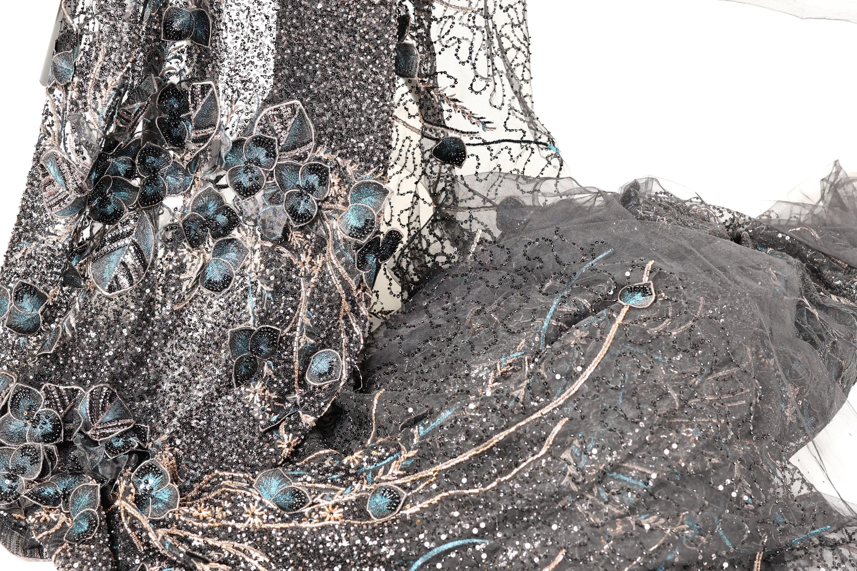 a close-up view of a fabric with intricate blue and black embroidered designs, including flowers and leaves. The fabric appears to be a sheer material, with a dark gray color and a textured pattern.