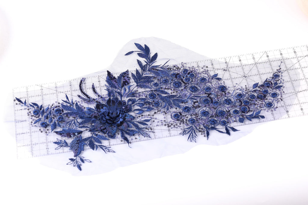 a close-up view of a piece of fabric with a floral pattern, including blue flowers and leaves. The fabric appears to be a lace or embroidered material, with a ruler visible in the background.