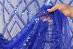A hand is holding up a piece of blue sequin fabric, showcasing its intricate and vibrant pattern.