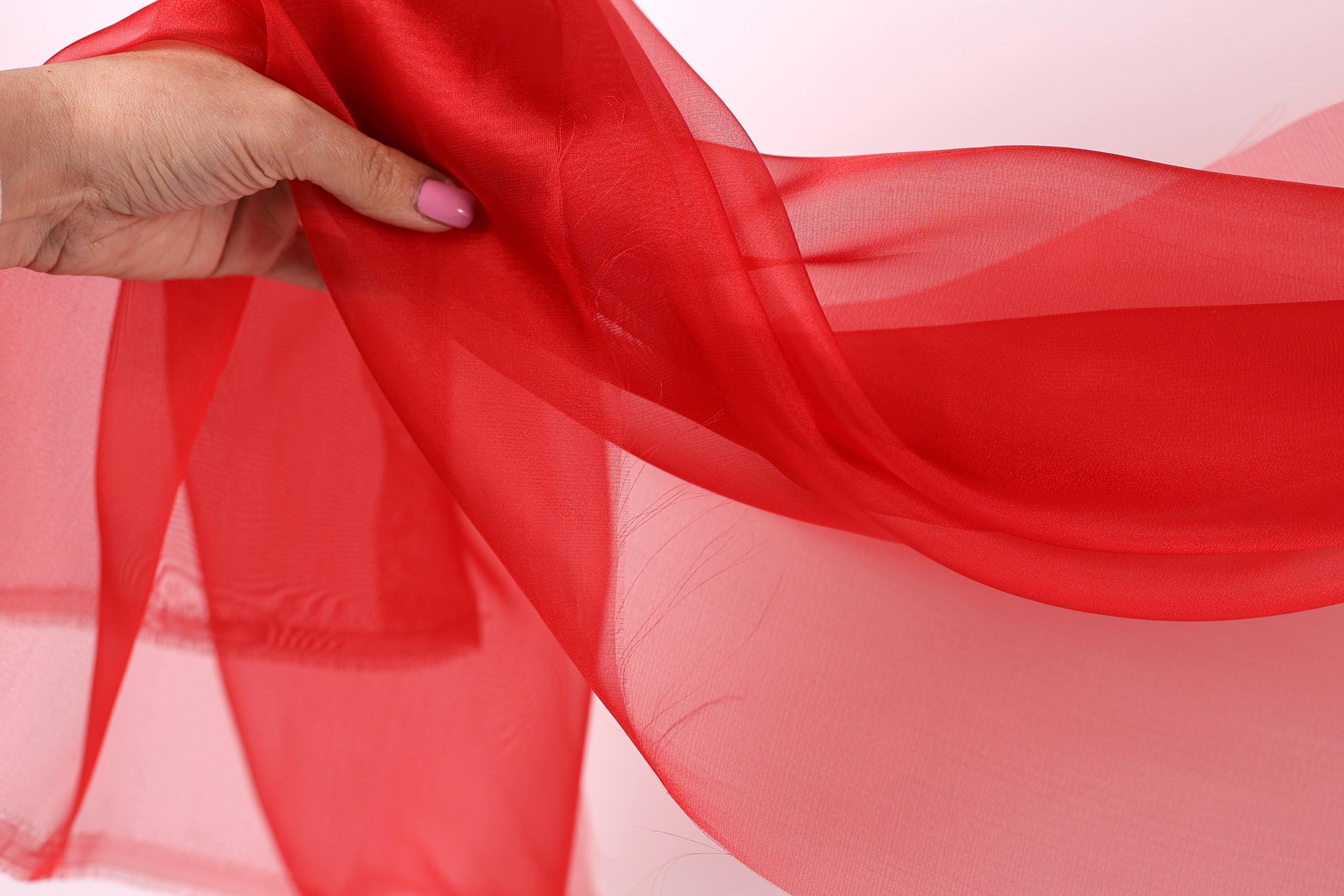 A hand holding a red fabric, possibly silk or chiffon, against a white background.